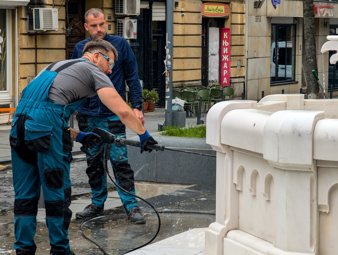 Two workers cleaning a fountain with a pressure washer in an urban setting.